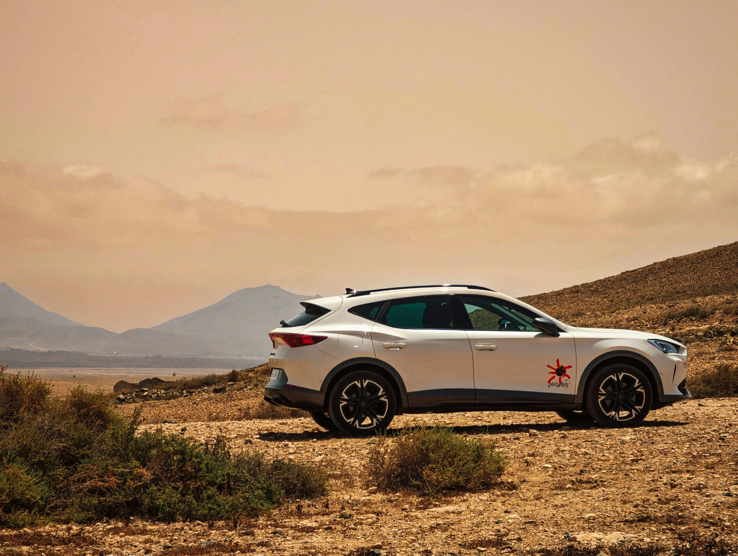 Sleek white car parked on rugged desert terrain with mountains in the background, under a warm sky.